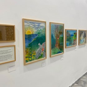 Eine Reihe gerahmter, farbenreicher Landschaftsbilder ist entlang einer wei&szlig;en Wand angeordnet. Die Werke zeigen unter anderem Meer, Sonne, Berge, B&auml;ume und fantasievolle Naturmotive.