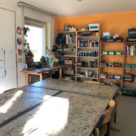 In einem Atelierraum steht zentral ein gro&szlig;er Arbeitstisch mit St&uuml;hlen. An einer Wand befinden sich Regale mit verschiedenen Materialien.