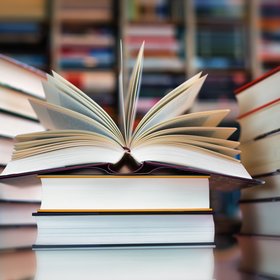 Drei Stapel von B&uuml;chern liegen auf einem Tisch. Auf dem mittleren liegt ein aufgeschlagenes Buch.